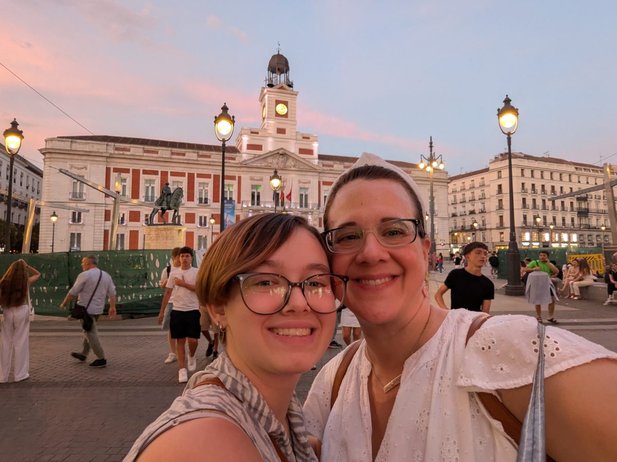 How to Avoid Common Travel Planning Mistakes in Your Next Vacation Two smiling women take a selfie in a busy plaza at sunset, with a statue and clock-tower building behind them.